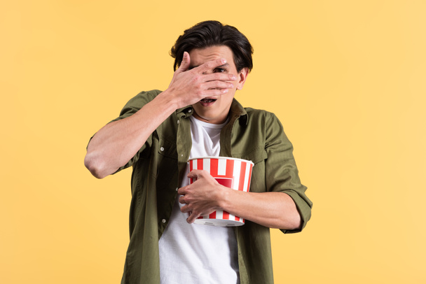 bange jongeman die horrorfilm kijkt, ogen sluit en emmer popcorn vasthoudt, geïsoleerd op geel - Foto, afbeelding