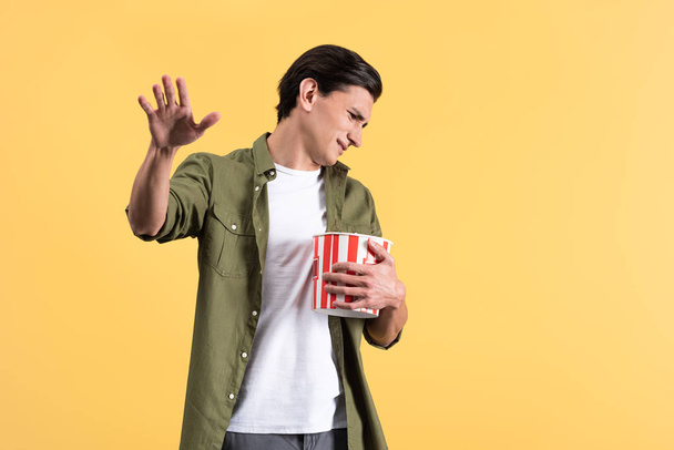 irritated young man watching movie with bucket of popcorn, isolated on yellow - Photo, image