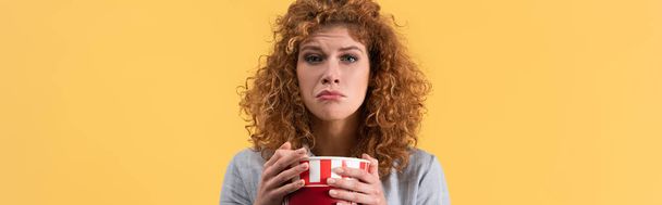 panoramic shot of sad girl watching movie with bucket of popcorn, isolated on yellow  - 写真・画像