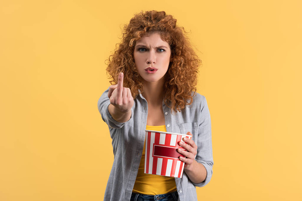 agressief meisje tonen middelvinger tijdens het kijken naar film met emmer popcorn, geïsoleerd op geel  - Foto, afbeelding