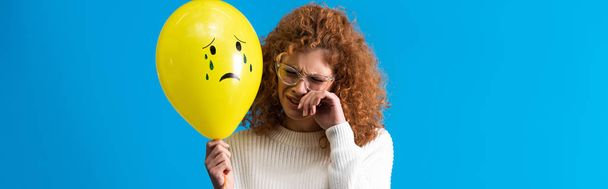 panoramic shot of upset redhead girl holding yellow balloon with crying face, isolated on blue - Zdjęcie, obraz