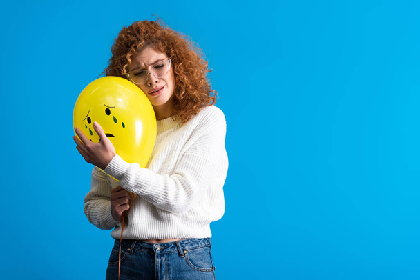 sad redhead girl holding yellow balloon with crying face, isolated on blue - Photo, Image