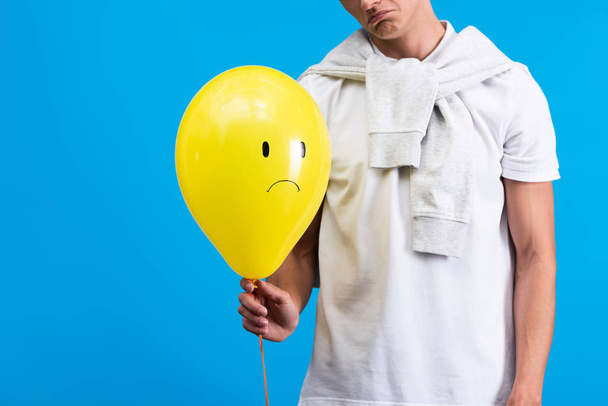 cropped view of sad man holding yellow upset balloon, isolated on blue - Foto, Imagem