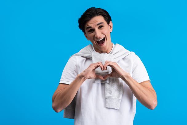 Porträt eines aufgeregten jungen Mannes, der Herzzeichen mit Händen macht, isoliert auf blau - Foto, Bild
