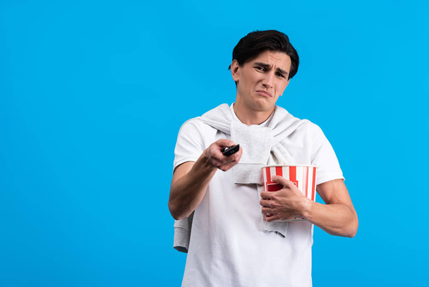 sad young man watching tv with remote controller and popcorn, isolated on blue - Foto, afbeelding