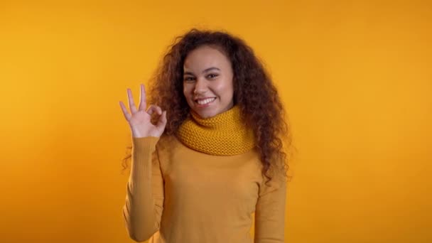 Positive girl making OK sign over yellow background and smiles to camera. Body language. Young curly woman in total warm look - Felvétel, videó