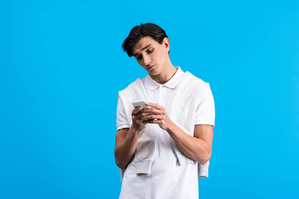 skeptical young man using smartphone, isolated on blue - 写真・画像