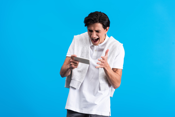 agressieve jongeman schreeuwen en met behulp van smartphone, geïsoleerd op blauw - Foto, afbeelding