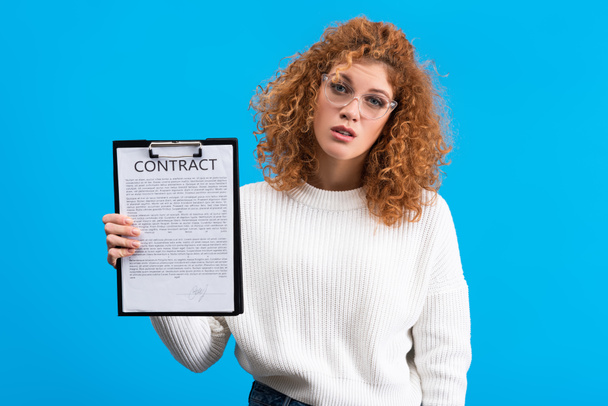 привлекательная скептически настроенная девушка держит планшет с контрактом, изолированный на голубом
 - Фото, изображение