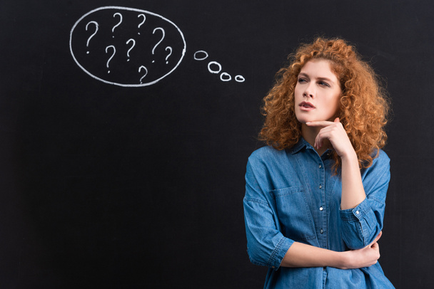 Nachdenkliche rothaarige junge Frau mit Fragezeichen in Gedankenblase auf Tafel - Foto, Bild
