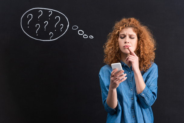 pensive woman using smartphone, question marks in thought bubble on blackboard behind  - 写真・画像