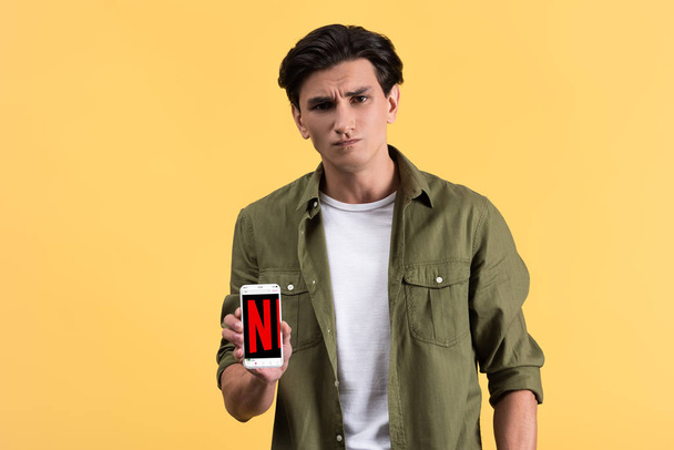 KYIV, UKRAINE - NOVEMBER 18, 2019: skeptical man showing smartphone with netflix app on screen, isolated on yellow  - Foto, Imagen