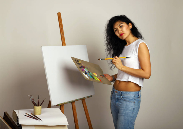 そのイーゼルの画家は絵を描く。筆塗りの魅力的な女の子。新しい絵画のための空白のキャンバスとブラシと塗料を持つアーティスト. - 写真・画像