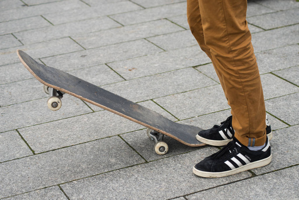patinador pernas vestindo tênis preto por Adidas esperando com skate bordo na rua
 - Foto, Imagem