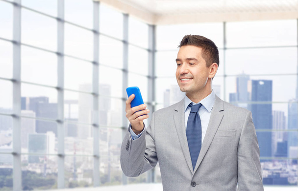 business, people and technology concept -happy businessman texting on smartphone over office room and window with city view background - Photo, Image