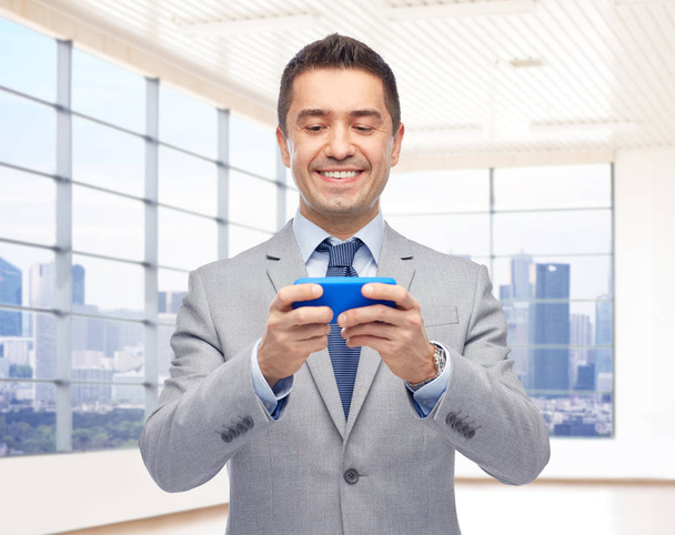 business, people and technology concept -happy businessman texting or playing game on smartphone over city office room window background - Φωτογραφία, εικόνα