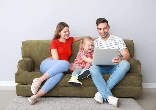 Happy young family with laptop sitting on sofa near light wall - Фото, изображение