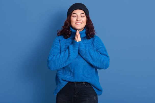 Image of cheerful adorable young lady standing with closed eyes isolated over blue background in studio, wearing dark blue sweater and hat, black trousers, smiling sincere. Emotions concept. - Foto, imagen