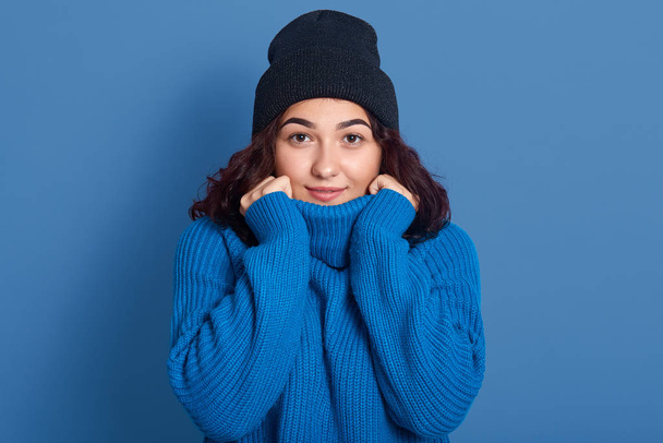 Closeup portrait of good looking magnetic charismatic young female looking directly at camera, touching blue sweater with hands, having peaceful facial expression, wearing blue sweater and hat. - Foto, Bild