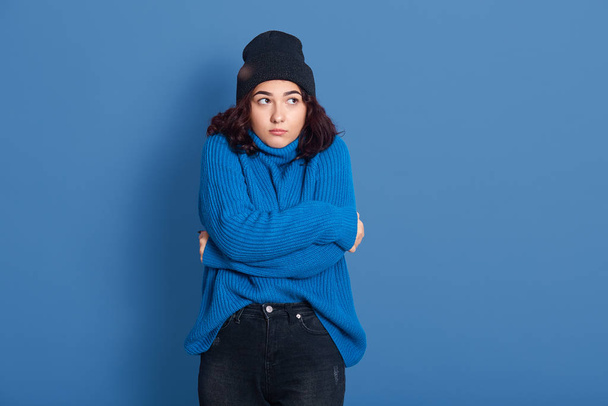 Horizontal studio picture of emotional upset female standing isolated over blue background, looking aside, hugging herself, feeling cold, winter concept, wearing warm sweater, hat and trousers. - Photo, Image