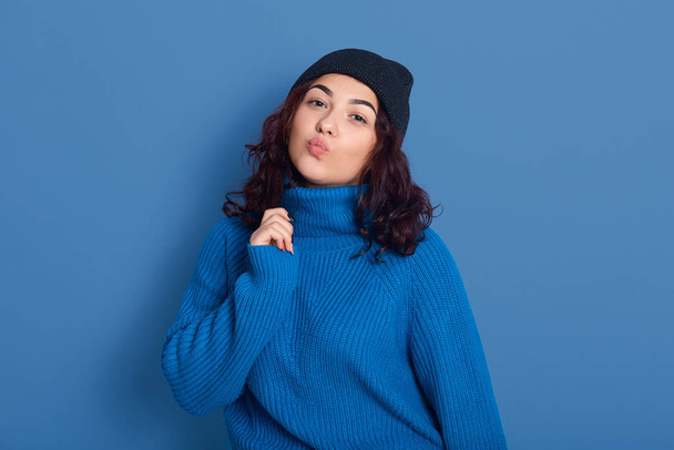 Indoor studio shot of good looking attractive cute young female raising one hand, putting lips together, kissing, having black curly hair, wearing dark blue sweater and hat. Emotions concept. - Foto, Bild