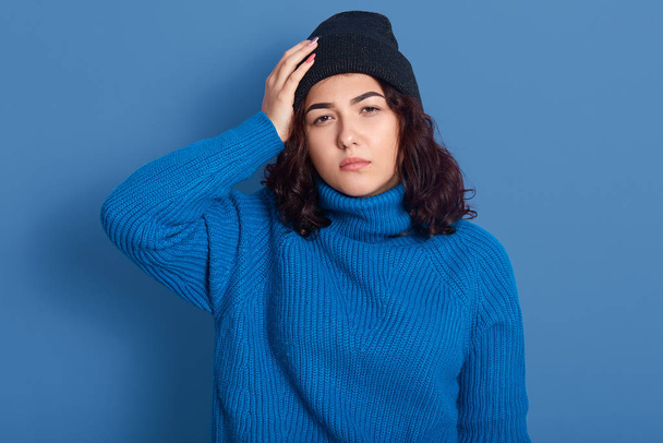 Portrait of thoughtful upset disappointed young lady with black curly hair, touching her head with hand, feeling unwell, having headache, standing isolated over phantom blue background in studio. - 写真・画像