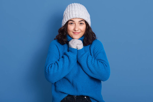 Image of beautiful caucasian darkhaired modest woman wearing warm cap and sweater, attractive model posing isolated over blue background in studio, keeps fists under chin, looking directly at camera. - Fotografie, Obrázek