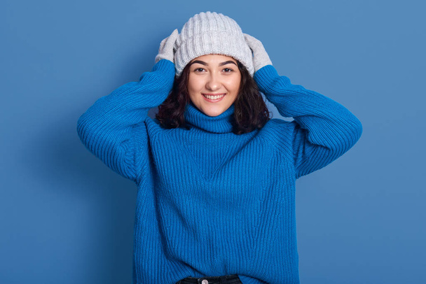 Portrait of happy young beautiful brunette girl smiling female, looking at camera, touching her head, posing isolated over blue background, girlwearing cap, mittens, sweater, lady expressing joy. - 写真・画像