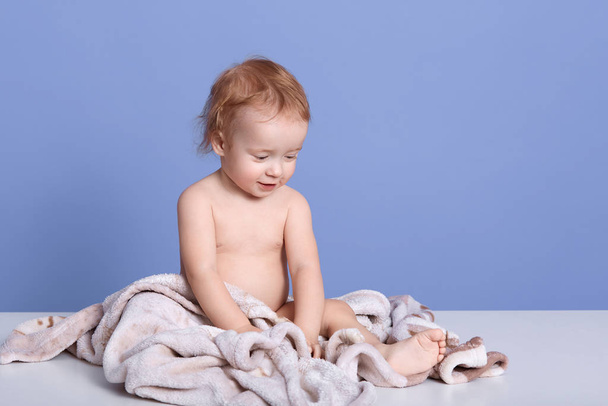Horizontal shot of charming baby boy sitting on towel isolated over blue background, adorable infant after bathing, nude cheeper playing and looking at hisbare leg with interest. Childhood concept. - Photo, Image