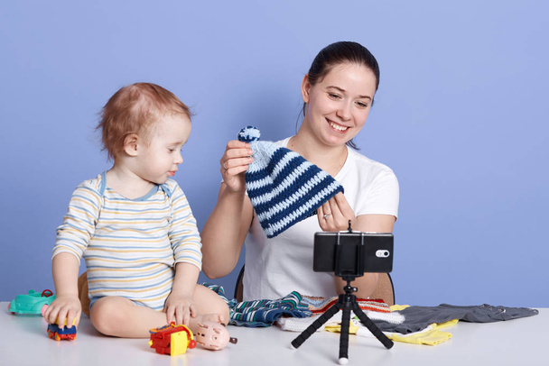 Close up portrait of contemporary young woman holding knitted cap while filming video for fashion and beauty channel against blue background, posing with her child isolated over blue background. - Fotoğraf, Görsel