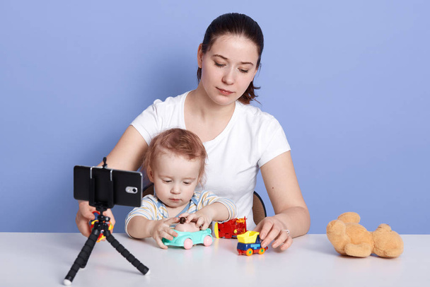 Mother and son playing with toys isolated over blue studio background, making content for her blog, infant being fond of car toy, looks concentrated and interested in playing. Blogging concept. - Fotografie, Obrázek