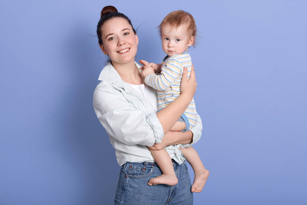 Indoor shot of happy family mother and child baby girl hugging kid, posing isolated over blue background, young female wearing shirt and jeans, looking smiling directly at camera. Family concept. - Photo, image