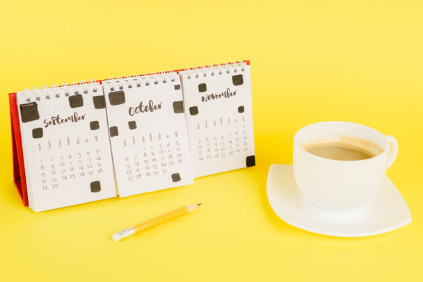 Cup of coffee near calendar and pencil on yellow background - Фото, зображення