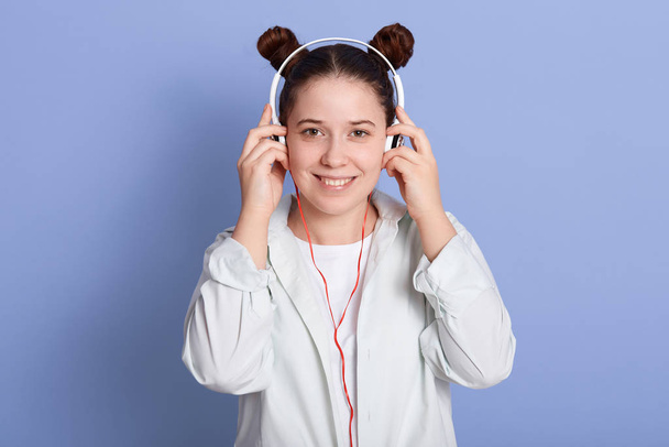 Image of beautiful girl with double buns hairstyle looking directly at camera while listening to music via headphones isolated over blue studio wall, captivating female wearing white clothing. - 写真・画像