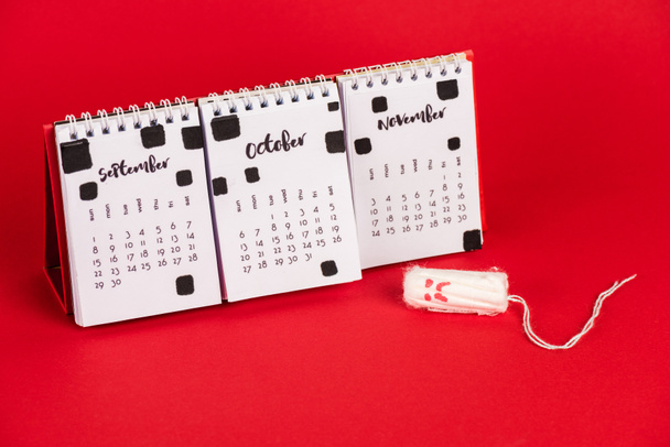 Calendar and hygienic tampon with sad face expression on red background - Photo, Image