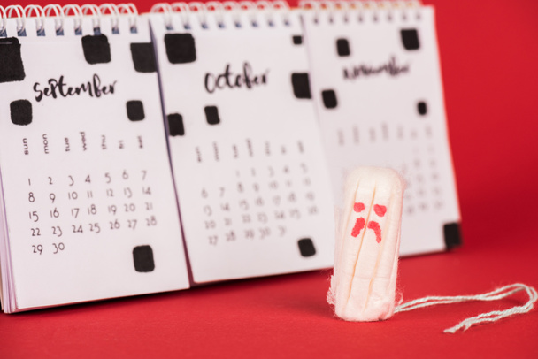 Selectieve focus van hygiënische tampon met droevige gezichtsuitdrukking nabij kalender op rode achtergrond - Foto, afbeelding