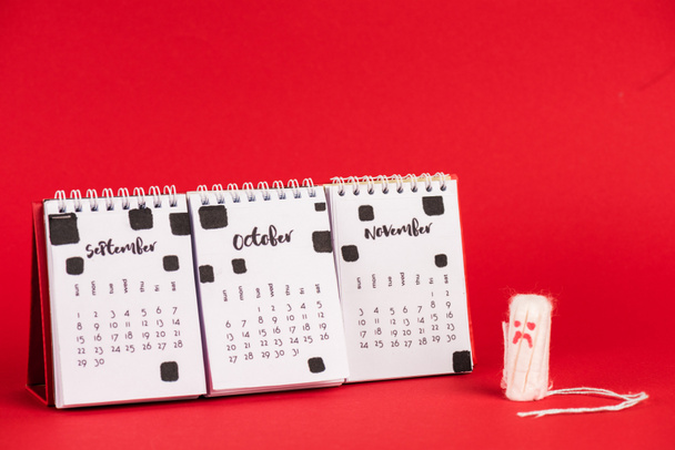Hygienic tampon with sad face expression near calendar with autumn months on red background - Photo, Image