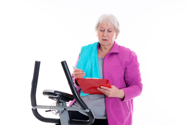 gray-haired female senior with clipboard train with fitness machine - Foto, Bild