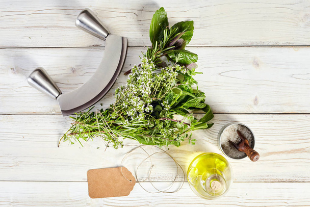 Mezzaluna knife, bouquet garni of assorted fresh culinary herbs tied in a bunch, olive oil and salt with a blank tag on an old painted white table with copy space - Фото, изображение