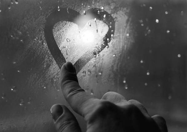 Καρδιά στο παράθυρο. Βαμμένο με ένα δάχτυλο σε θολό γυαλί κατά τη διάρκεια της βροχής. Φωτεινό bokeh από μια λάμπα του δρόμου. - Φωτογραφία, εικόνα