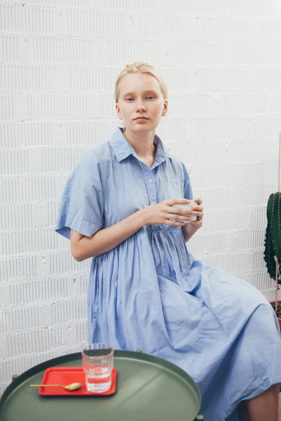Retrato de una hermosa mujer joven en vestido azul que está bebiendo café especial en un café moderno con un mínimo interior de estilo escandinavo. Luz natural, pared de ladrillos blancos en el fondo
. - Foto, imagen