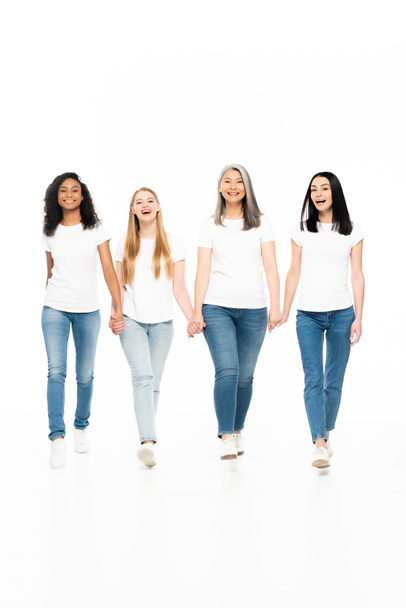 szczęśliwe wielokulturowe kobiety w dżinsach jeansowych chodzenie i trzymanie się za ręce odizolowane na białym  - Zdjęcie, obraz