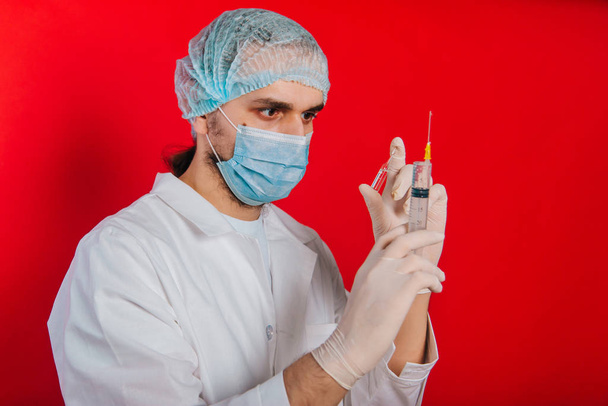 Der Arzt hält eine Spritze und eine Ampulle mit einem Impfstoff in der Hand. ein junger Mann in weißem Mantel, mit medizinischer Maske und Handschuhen auf rotem Hintergrund. - Foto, Bild