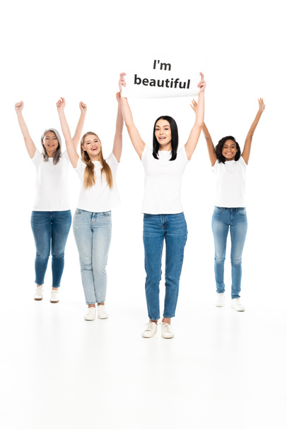 happy multicultural women in denim jeans near placard with i`m beautiful lettering isolated on white  - 写真・画像