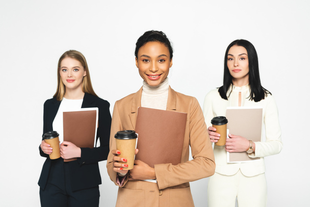 happy multicultural businesswomen holding folders and paper cups isolated on white  - Photo, Image