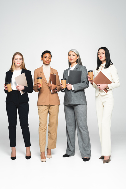 four multicultural businesswomen holding folders and paper cups while standing on white  - Foto, Bild
