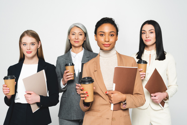 four attractive and multicultural businesswomen holding folders and paper cups isolated on white  - Φωτογραφία, εικόνα
