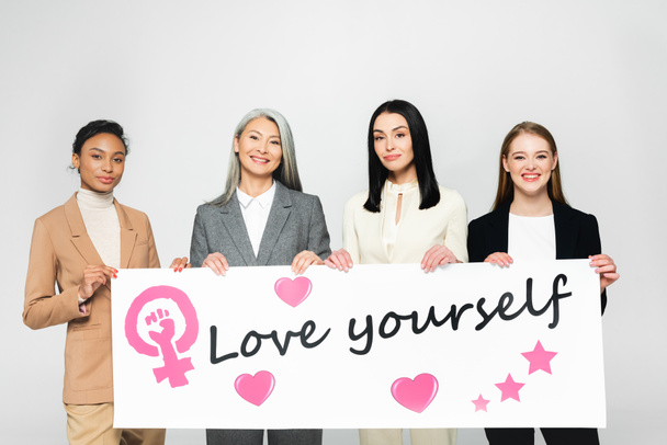 glückliche multikulturelle Geschäftsfrauen, die ein Poster mit dem Schriftzug "Liebe dich selbst" in der Hand halten  - Foto, Bild