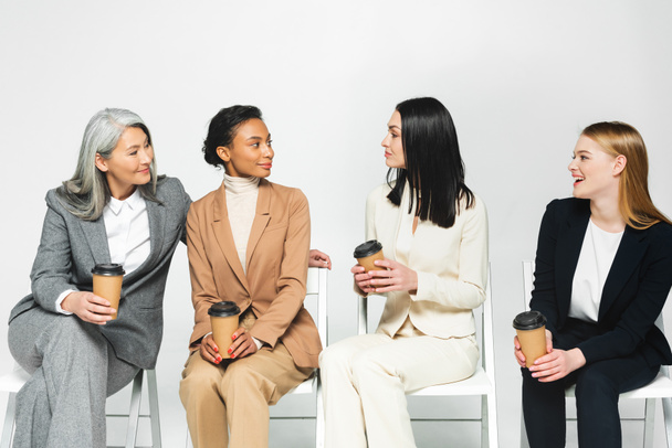 mujeres de negocios multiculturales felices en trajes sentados en sillas y sosteniendo vasos de papel aislados en blanco
  - Foto, Imagen