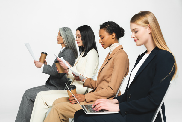 side view of multicultural businesswomen sitting on chairs with paper cups and laptop on white  - Photo, Image